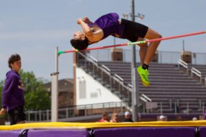 Male high jumper in purple uniform clears a pink bar over purple and yellow padded mats, with spectators and bleachers in the background.