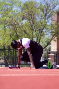 Athlete crouched at the starting line on a red track, holding a purple baton with green trees in the background.