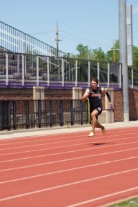 Male sprinter in black Coyotes jersey (#5) running on an outdoor red track beside stadium bleachers.