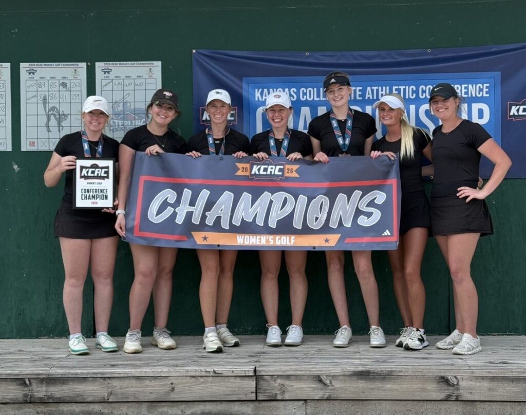 Seven female golfers in black outfits pose on a wooden platform, holding a KCAC Champions banner and trophy, with scoreboards and a blue banner in the background indicating Women's Golf.