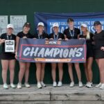 Seven female golfers in black outfits pose on a wooden platform, holding a KCAC Champions banner and trophy, with scoreboards and a blue banner in the background indicating Women's Golf.