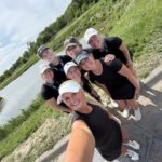 Seven women in black athletic outfits taking a selfie by a pond on a sunny day, smiling with green grass and trees in the background.