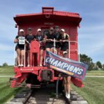 Women’s golf team standing on a red train caboose, holding a plaque and a large banner that reads 'Champions' outdoors on a sunny day.
