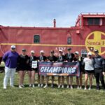Women’s golf team posing with medals and a 'Champions' banner in front of a red Santa Fe locomotive on a sunny day.