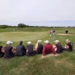 Group of women golfers sitting on the grass watching players near the green on a sunny course.
