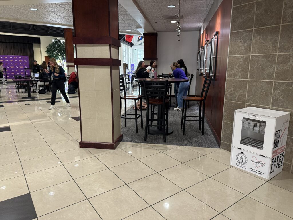 People socialize at high-top tables in a bright campus lobby; a purple event backdrop is visible on the left. A white donation/food kiosk sits at the right foreground.
