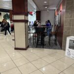 People socialize at high-top tables in a bright campus lobby; a purple event backdrop is visible on the left. A white donation/food kiosk sits at the right foreground.