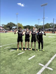 Four male athletes in black Coyotes uniforms stand together on a sunny football field, making rock-on hand gestures with smiles.