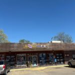 Front of a brick storefront with a wooden awning and a yellow sign showing 'KW' above the entrance, blue sky in the background, and posters in the windows. Parking lot with a couple of cars visible in front.