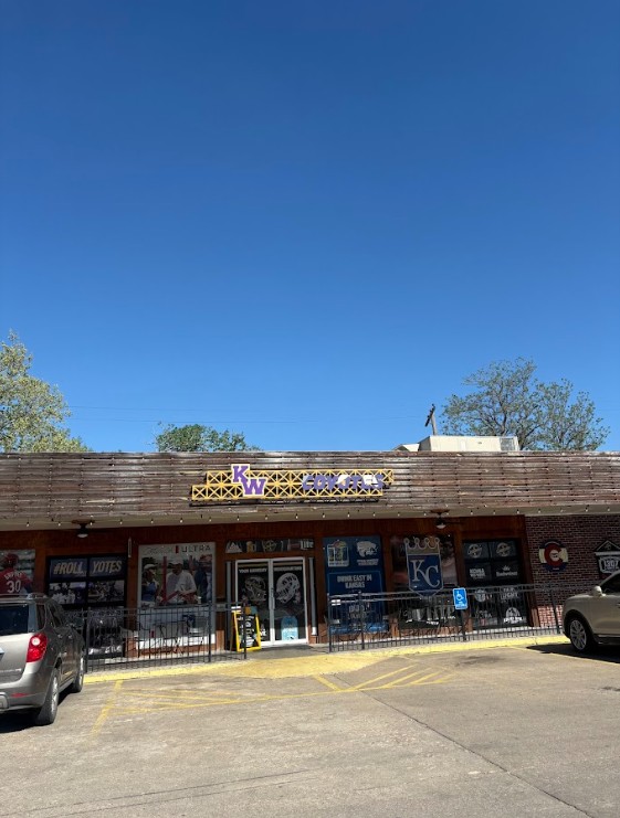 Front of a brick storefront with a wooden awning and a yellow sign showing 'KW' above the entrance, blue sky in the background, and posters in the windows. Parking lot with a couple of cars visible in front.