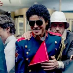 Elvis-inspired performer in a blue military jacket with gold buttons and red sash, wearing sunglasses, surrounded by a crowd.