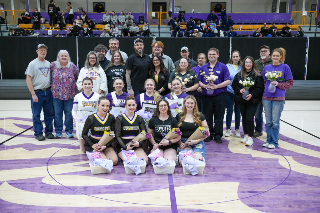 Group of cheerleaders in purple uniforms posing with bouquets and fans in a gymnasium backdrop.