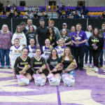 Group of cheerleaders in purple uniforms posing with bouquets and fans in a gymnasium backdrop.