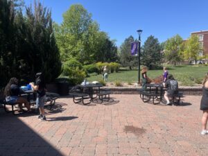 Group of people sitting and standing around round metal tables in a brick-paved campus courtyard on a sunny day, with trees and a blue sky in the background.