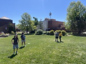 Children outside on a sunny campus lawn with green grass, trees, and brick buildings in the background.