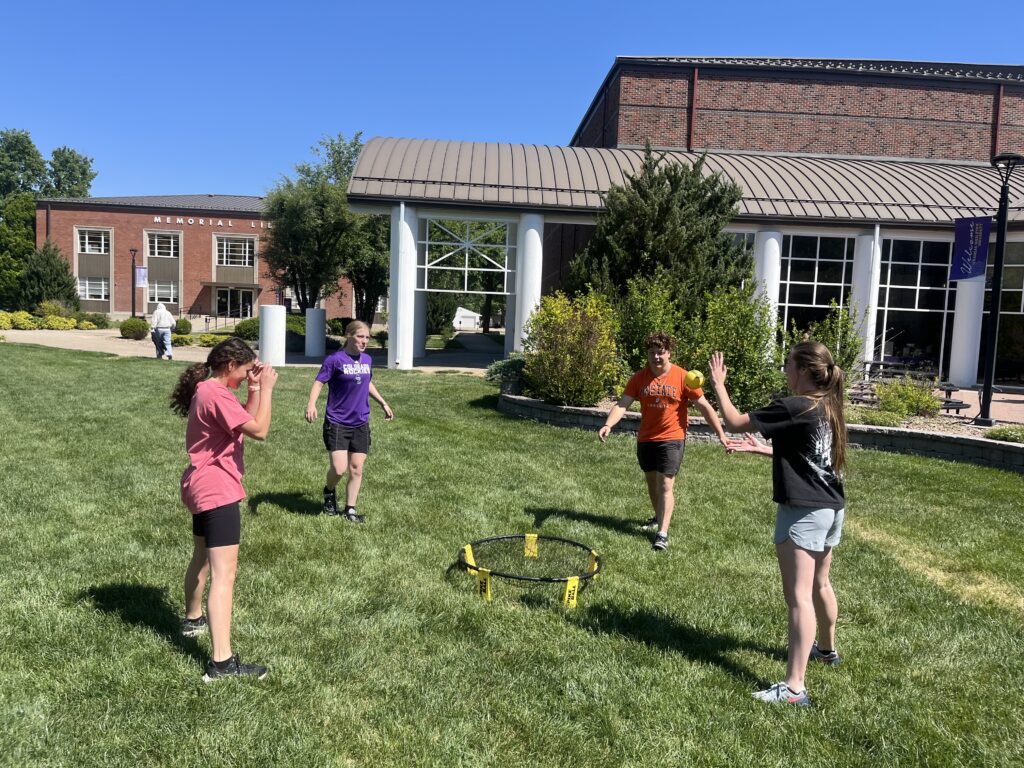 Group of kids playing Spikeball on a sunny campus lawn with brick buildings in the background.