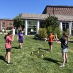 Group of kids playing Spikeball on a sunny campus lawn with brick buildings in the background.