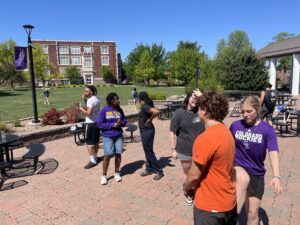 Group of students chatting in a sunny campus courtyard with a red-brick building, trees, and outdoor seating behind them.