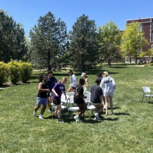 Group of students outdoors on a sunny campus lawn, gathered around white folding chairs with trees and a brick building in the background.