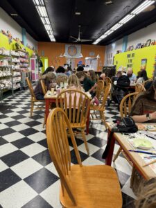 Busy craft shop cafe; people sit at wooden tables painting ceramics in a colorful store.