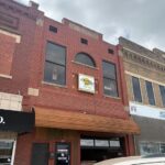 Two-story brick storefronts on a city street; central building has an awning and a sign, with a car in the foreground.
