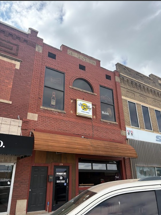 Two-story brick storefronts on a city street; central building has an awning and a sign, with a car in the foreground.