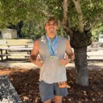 Man posing outdoors under a leafy tree, showing two race medals on a blue lanyard and giving a thumbs-up gesture. He wears a gray sleeveless shirt, dark shorts, and sunglasses on his head with a race bib visible on his side.